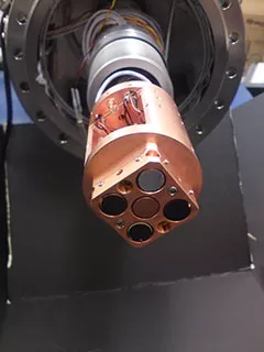 Within the space environment testing chamber, the interchangeable sample pedestal holds a central Faraday cup and four slots for 10mm copper cylinders, each with a different material specimen mounted with Master Bond EP21TDCS-LO conductive epoxy.[1]